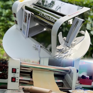 Peut inclure: Machine à pâtes blanche avec rouleaux en acier inoxydable et une feuille de pâte. La machine a un interrupteur d'alimentation et une mesure de 40 cm. Un rouleau à pâtisserie en bois et une feuille de pâte sont également visibles.
