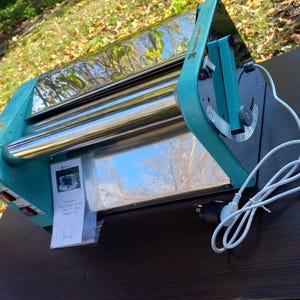 May include: A teal and chrome electric dough roller machine featuring a side-mounted adjustment handle, a power switch, and a white power cord. A small plastic bag containing a printed instruction manual is attached to the front of the device.