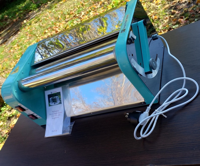 May include: A teal and chrome electric dough roller machine featuring a side-mounted adjustment handle, a power switch, and a white power cord. A small plastic bag containing a printed instruction manual is attached to the front of the device.