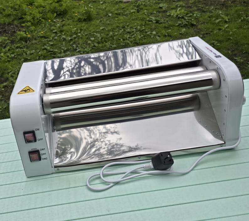 May include: A white electric food warmer with two horizontal polished stainless steel rollers. The unit features a side control panel with two black rocker switches labeled ON/OFF and SPEED ON1/OFF/ON2, a yellow warning icon, and a white power cord.