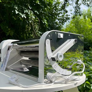 May include: A white and chrome electric pasta machine featuring a side-mounted adjustment lever with printed markings for thickness settings ranging from 1/32 inches to 13/16 inches, a power switch, and a white power cord with a standard plug.