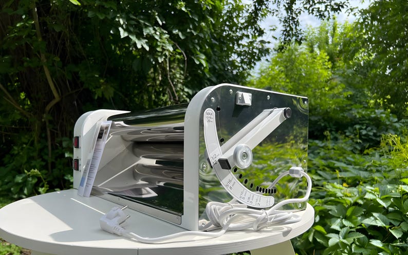 May include: A white and chrome electric pasta machine featuring a side-mounted adjustment lever with printed markings for thickness settings ranging from 1/32 inches to 13/16 inches, a power switch, and a white power cord with a standard plug.