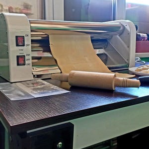 May include: A commercial pasta machine in beige and silver, processing a sheet of dough. A wooden rolling pin is on the dark brown countertop. The machine has two red power switches and a clear plastic packet.