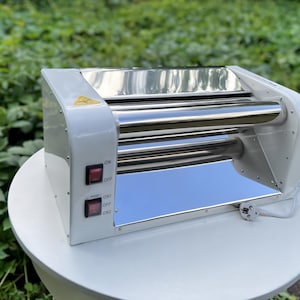 以下が含まれることがあります： 白とシルバーのラミネーター機。赤色の電源ボタンが2つあり、「ON」と「OFF」と表示されています。機械は光沢のある反射面と黄色の三角形の警告記号を備えています。白いテーブルの上に置かれています。