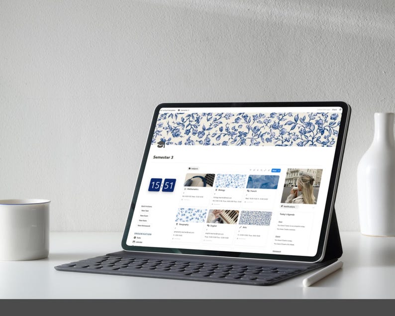 Peut inclure: Une tablette affiche une interface num&eacute;rique avec un motif floral bleu. L'&eacute;cran montre un calendrier et des ic&ocirc;nes d'applications. Une tasse blanche, un vase blanc et un stylet sont &eacute;galement visibles.