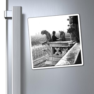 May include: Black and white photograph of a gargoyle statue on a building overlooking a city. Two people are sitting on the ledge of the building, looking out at the city.