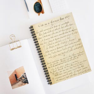May include: A spiral-bound notebook featuring a vintage-style cover with the handwritten text Ode on a Grecian Urn 1819. The notebook is accompanied by a gold metal binder clip, a pair of brown-rimmed sunglasses, and a small square photograph.