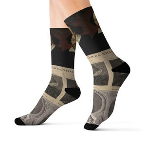 May include: Black and beige socks featuring a portrait of William Shakespeare and the words "Shakespeare". The socks have a black top and toe, with a beige and black design of a portrait of Shakespeare.