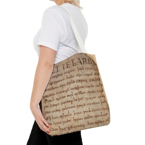 May include: A beige tote bag with a white strap. The bag features a vintage-style print of an old document with text in an old English font. The text reads "ET TE LARD 129 mzan dazum peod cynniza stpumon huda cepelimz elle Emedon. oft feild (ceems scerber Preacum monezu megum meodo recla off real srode coul syddan cequere pe fea scenye funden he þær poppe seba pesx under polemum pesid myndin pala od him aghpyle papa jomb firrendra eq hpon pade hypan scolde gombar yldan pper zod cynmz. dem ετfέμα μας Cros some".