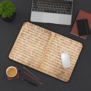 Puede incluir: Una alfombrilla de escritorio rectangular con un diseño impreso de un manuscrito antiguo abierto, con texto en tinta marrón oscuro sobre un fondo de pergamino beige. El texto comienza con las letras grandes y estilizadas "HÆT PE GARD". Un ratón blanco descansa sobre la alfombrilla.