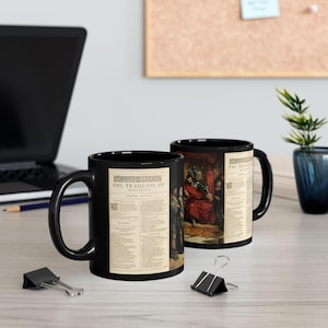 May include: Two black ceramic mugs with the text "THE TRAGEDIE OF MACBETH" printed on them. One mug features a scene from the play, while the other displays a page of text. The mugs are on a light-colored surface.