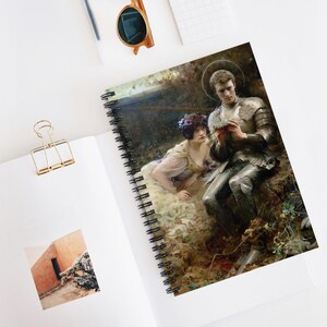 May include: A spiral-bound notebook featuring a reproduction of a painting depicting a person in medieval armor sitting with a person wearing a floral crown in a forest setting. The notebook is open, showing a small square photo of a building.