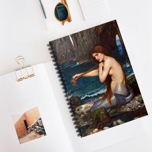 May include: A spiral-bound notebook featuring a reproduction of John William Waterhouse's painting of a mermaid with long red hair sitting on a rocky shore by the sea, holding a shell, with a black metal coil binding along the left side.