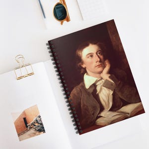 May include: Spiral-bound notebook featuring a portrait of a man in a brown jacket and white shirt, resting his chin on his hand. The notebook is open, revealing the portrait and a black spiral binding. The background is a dark maroon color.
