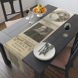 May include: A beige table runner featuring a vintage-style print of William Shakespeare's Hamlet title page, including the text The Tragical Historie of Hamlet, Prince of Denmarke, and a portrait illustration of Shakespeare, laid flat on a dark table.