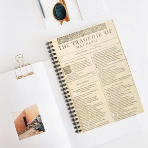 May include: A spiral-bound notebook with a vintage-style cover featuring the title "The Tragedie of Macbeth." The pages display printed text. A gold-colored paper clip and sunglasses are also visible.