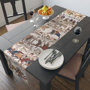 May include: A long table runner featuring a detailed, multi-paneled historical narrative print with figures, horses, and Latin text in muted earth tones of beige, brown, blue, and red, draped across a dark wooden dining table with place settings.