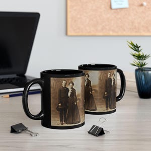 May include: Two black coffee mugs with a vintage photograph of a couple in formal attire. The photo shows a man and a woman standing side-by-side, both wearing hats. The photo is in sepia tones.