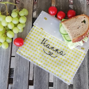 Könnte beinhalten: Ein wiederverwendbarer Lebensmittelverpackung aus gelbem und weißem Karo mit dem Namen "Hanna" in schwarzer Kursivschrift mit einem Herzen. Die Verpackung ist über ein Sandwich mit Salat, Gurke und Brot gefaltet.