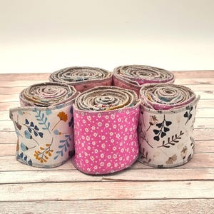 Könnte beinhalten: Sechs Stoffrollen mit floralen Mustern in Rosa, Blau und Gelb. Die Rollen sind auf einer hellen Holzoberfläche angeordnet, wodurch die verschiedenen Designs und Texturen des Stoffes hervorgehoben werden. Jede Rolle hat einen Durchmesser von etwa 10 cm.