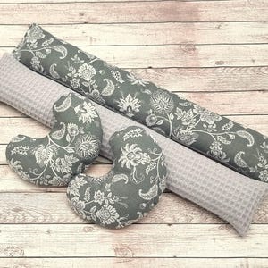 Peut inclure: Plusieurs coussins gris et blancs à motifs floraux et un long traversin. Les coussins sont de formes variées, dont des croissants et des rectangles, et sont disposés sur une surface en bois. Le tissu présente un motif floral détaillé.