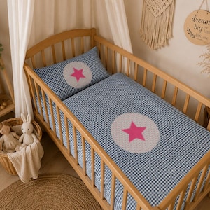 Puede incluir: Una cuna de madera con ropa de cama de cuadros azules y blancos. La ropa de cama presenta un gran círculo blanco con una estrella rosa en el centro. Una almohada a juego también está en la cuna. Una pequeña cesta de mimbre con animales de peluche está cerca.