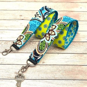 Op de afbeelding: Een turquoise en gele gepatroneerde stoffen lanyard met twee zilveren sleutelhangers. De lanyard is omkeerbaar en heeft een ander patroon aan elke kant.