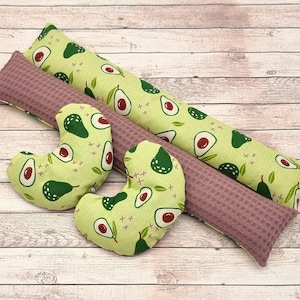 Könnte beinhalten: Eine Sammlung von Wohnaccessoires zum Thema Avocado. Enthält zwei lange, rechteckige Kissen mit hellgrünem Avocado-Aufdruck, ein mauvefarbenes Kissen mit Waffelstruktur und zwei kissenförmige Avocados. Der Avocado-Aufdruck zeigt grüne Avocados mit roten Kernen.