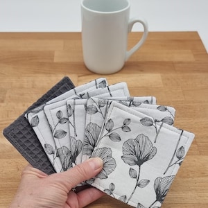 Può includere: Un set di sei sottobicchieri quadrati. Cinque sono bianchi con un motivo floreale nero, e uno è grigio scuro con una trama a nido d'ape. Una tazza bianca è sullo sfondo su una superficie di legno.