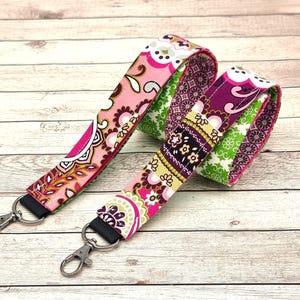 Può includere: Due cordini in tessuto con ganci girevoli. Uno presenta un motivo floreale rosa, bianco e giallo. L'altro ha un design floreale viola, verde e giallo. Entrambi i cordini hanno tessuto nero sul retro e ganci girevoli color argento.