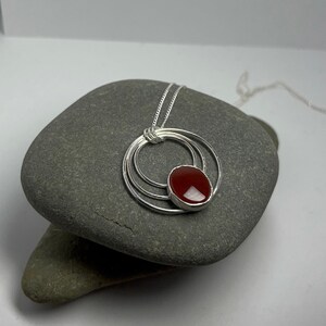 Puede incluir: Un collar de plata con un colgante de piedra de cornalina roja. El colgante está rodeado por tres anillos de plata.
