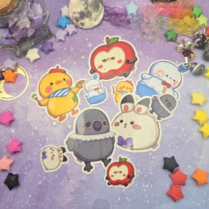 May include: A selection of vibrant stickers showcasing cartoon characters. The stickers depict a yellow chick with a blue scarf, a red apple with a grumpy expression, a white bunny with a purple bow, and a grey bird.