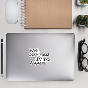 May include: A laptop sticker with the text "Well, look what TJMaxx dragged in." is displayed on a laptop. A white computer mouse, a spiral-bound notebook, a pen, glasses, and small potted plants are also visible.