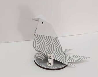 May include: A white origami bird with a gray and white patterned design, standing on a round wooden base. The bird has a small black dot for an eye and a white tag with the Japanese word "モミジ" written on it.