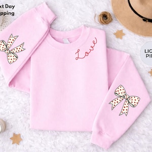 Puede incluir: Sudadera rosa claro con la palabra "Love" bordada en rojo. Las mangas presentan un diseño de lazo con corazones rojos. El texto "Next Day Shipping" es visible en la esquina superior izquierda, y "LIGHT PINK" en la esquina superior derecha.
