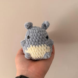 May include: A small, round, gray crocheted plush toy with a soft, textured yarn finish. The creature features two small upright ears, two black bead eyes, and a cream-colored patch on its belly. The design is compact and rounded in shape.