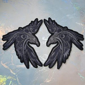 Könnte beinhalten: Zwei schwarze gestickte Raben-Patches mit aufwendigen Details. Die Raben schauen sich mit ausgebreiteten Flügeln an.