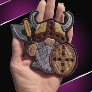 May include: A brown and gray embroidered patch of a Viking gnome wearing a horned helmet and holding a shield and axe.