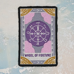 Może przedstawiać: Haftowana naszywka przedstawiająca kartę tarota Koło Fortuny. Prostokątna naszywka ma czarną obwódkę. Projekt zawiera fioletowe koło, różowe chmury i złote akcenty z napisem "WHEEL OF FORTUNE".