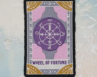 May include: Embroidered patch featuring the Wheel of Fortune tarot card design. The patch has a rectangular shape with a black border. The design includes a purple wheel, pink clouds, and gold accents, with the words "WHEEL OF FORTUNE".