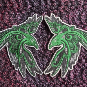Könnte beinhalten: Zwei gestickte schwarze und grüne Aufnäher mit einem stilisierten Vogel mit ausgebreiteten Flügeln. Der Vogel hat einen grünen Umriss und komplizierte Details.