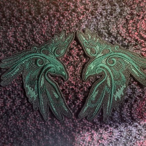 Könnte beinhalten: Zwei grüne gestickte Aufnäher mit stilisierten Vögeln und filigranen Details. Die Aufnäher befinden sich auf einem strukturierten lila und schwarzen Hintergrund.