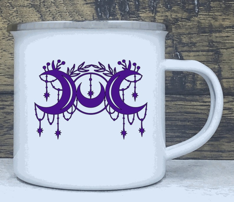 May include: White enamel mug with a purple triple moon design. The moon phases are surrounded by a floral design and dangling stars.