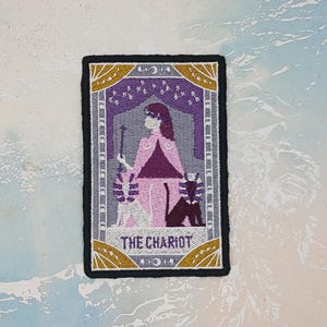 以下が含まれることがあります： タロットカードのデザインが施された刺繍パッチ。パッチは黒い縁取りで、ピンクと紫の人物が杖を持ち、「THE CHARIOT」の文字が下部にあります。金とグレーのアクセントがデザインを完成させています。