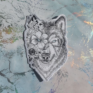 May include: A black and white embroidered patch of a wolf's face with flowers covering one eye. The wolf's fur is detailed with shading and the flowers are in a variety of shapes and sizes.