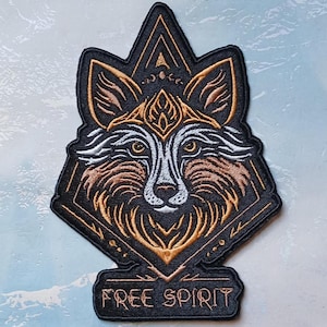 May include: Embroidered patch featuring a stylized wolf head with geometric details in black, brown, white, and gold. The patch has the words "FREE SPIRIT" at the bottom. The patch is diamond-shaped with a triangular top.