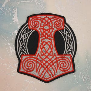 May include: An embroidered patch featuring a red and silver Mjolnir hammer design with intricate knotwork patterns. The central hammer shape is outlined in black, set against a black background, and surrounded by a circular silver Celtic knot border.