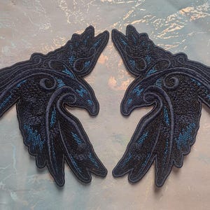 May include: Two black embroidered raven patches with blue accents. The ravens are facing each other with their wings spread out.
