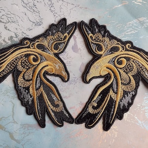 Könnte beinhalten: Zwei schwarze gestickte Aufnäher mit goldfarbenem Garn, die stilisierte Vögel mit komplizierten Designs darstellen. Die Vögel sind in entgegengesetzte Richtungen gerichtet.