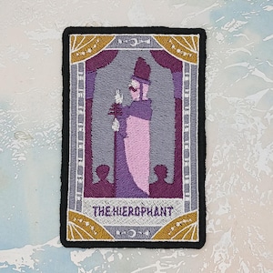 Puede incluir: Parche bordado con un diseño de carta del tarot, "THE HIEROPHANT." El parche tiene un borde negro, con una imagen central de una figura vestida con túnica morada y rosa, sosteniendo un bastón. El fondo es gris con detalles dorados.